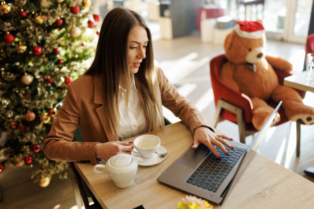 A woman sits at her laptop in a café. There is a Christmas tree behind her and other festive decorations. 