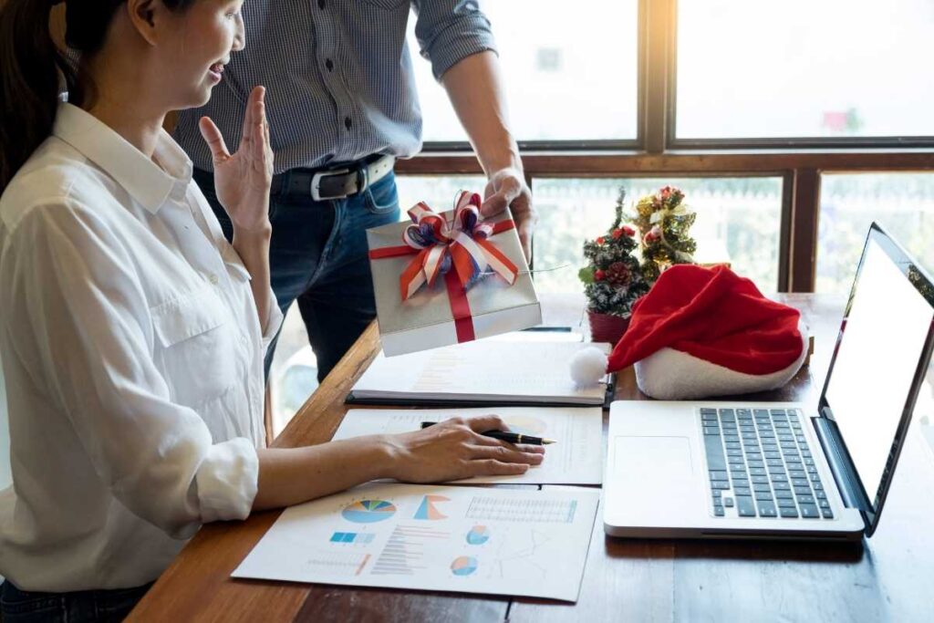 a female office worker is handed a wrapped christmas gift whilst sat at her desk working. There is a small christmas tree and santa hat. 