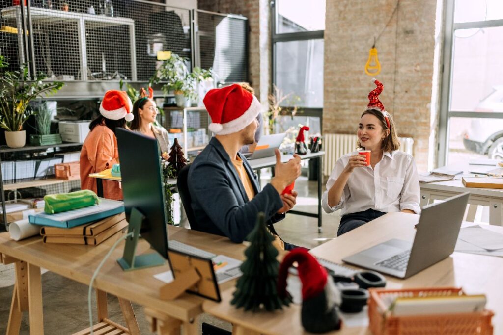 two colleagues sat chatting to each other in an office, both wearing festive headwear and drinking out of red cups.