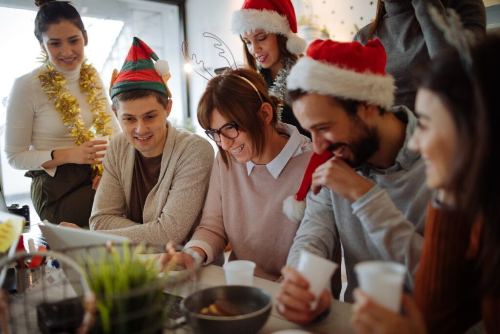 group of office workers stood smiling over a laptop. they are all wearing Christmas hats, reindeer ears and tinsel