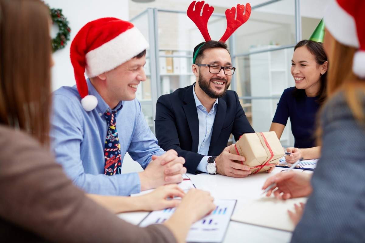 A man holds his Christmas corporate gift in a meeting. His team wear santa hats and reindeer antlers