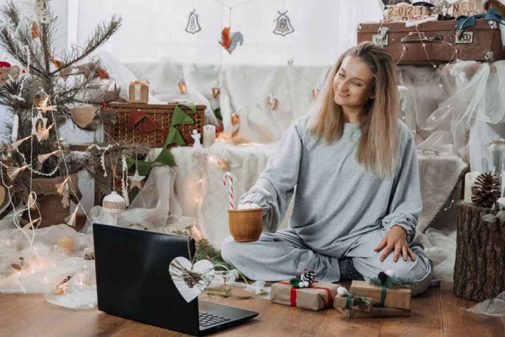 a woman sits on the floor surrounded by christmas decor. She is drinking hot chocolate and working on her laptop. 