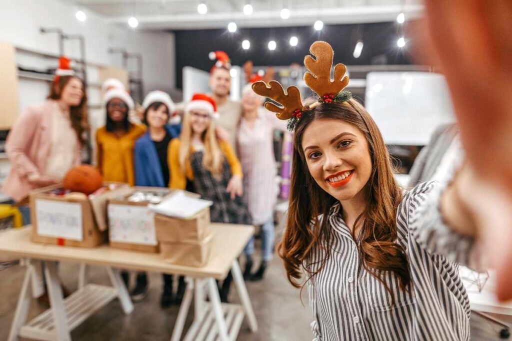 a woman takes a selfie at a Christmas workplace charity event, she is wearing reindeer ears and her team are behind her wearing Santa hats and smiling.