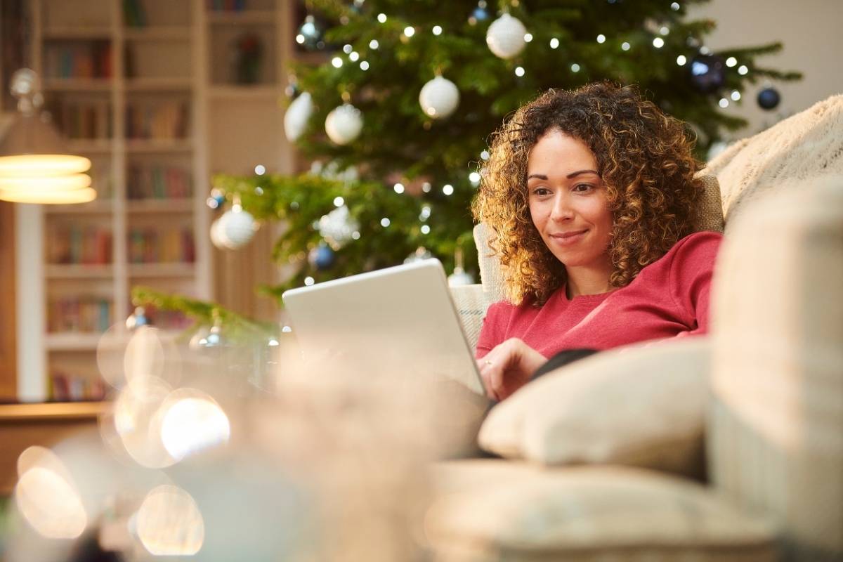 smiling woman sits on her sofa whilst using her laptop. there is a christmas tree behind her