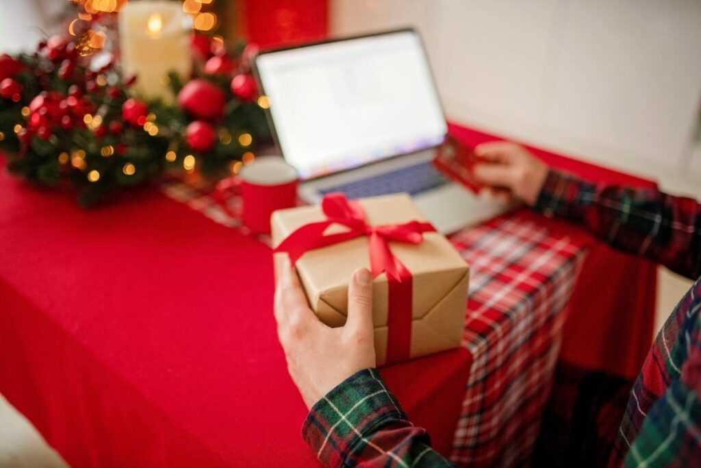 a man sits at a christmas table and holds a wrapped gift in one hand and his card in the other. he sits in front of a laptop and a festive table wreath. 