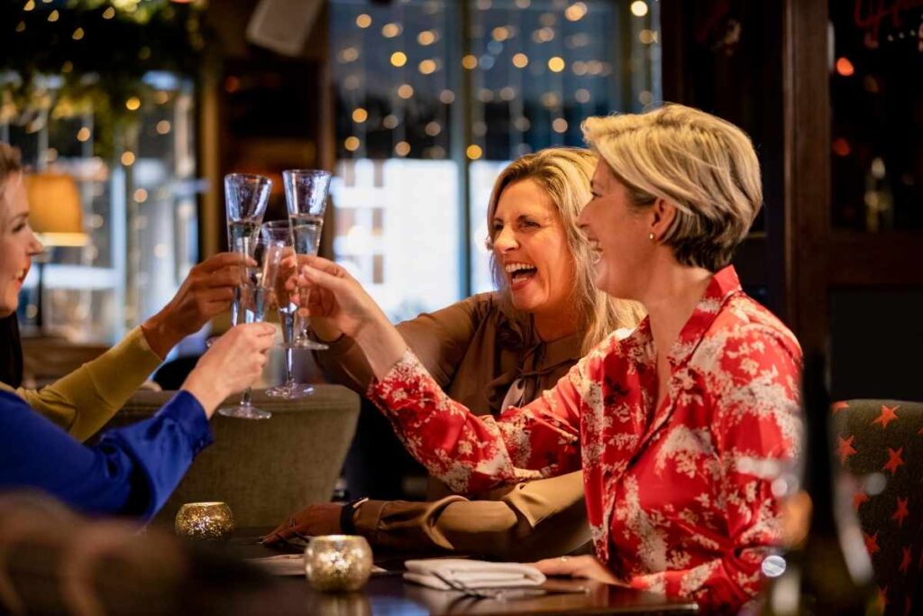 three woman cheers three glasses of prosecco and smile together.  there are Christmas lights in the background. 