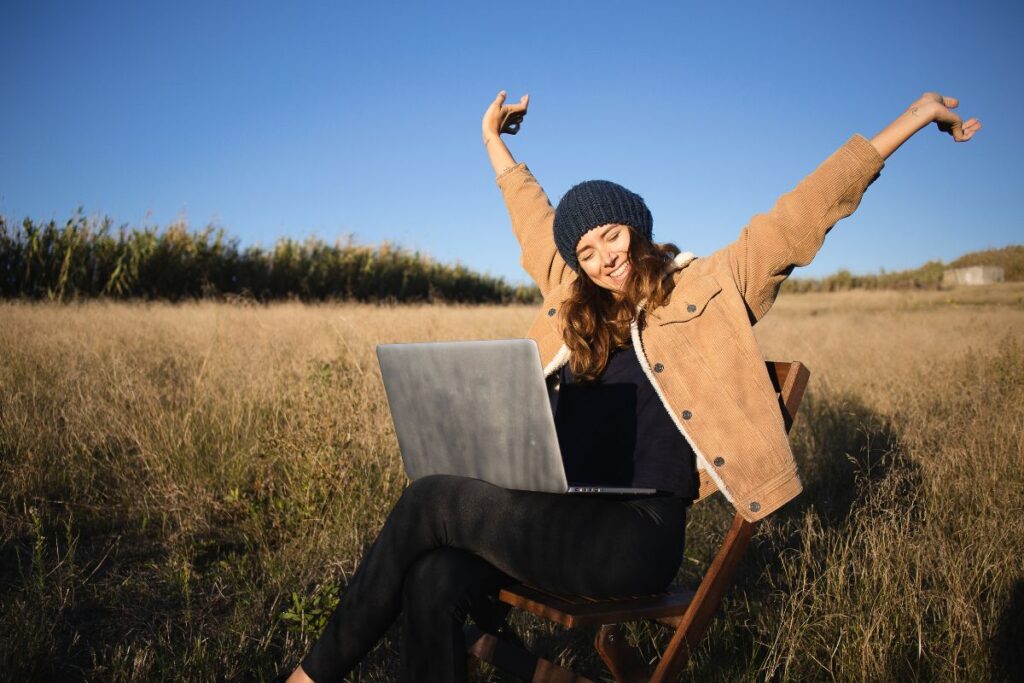 a woman stretches her arms up as she sits on a chair in a field. she has a laptop on her lap.