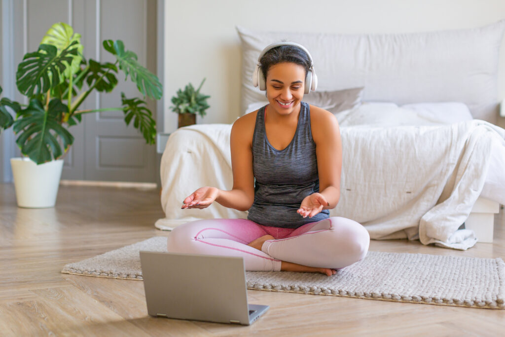 Woman sitting cross-legged on floor mat in front of her laptop, wearing workout clothes and headphones. 