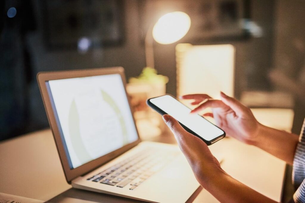 Hands scroll on a smart phone. They are sat in front of a laptop, with a desk lamp in the background. 