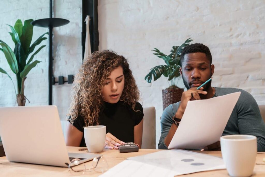 two professionals sit at a desk with papers and a laptop. They are in a discussion whilst using a calculator. There are coffee cups on the desk and plants in the background.