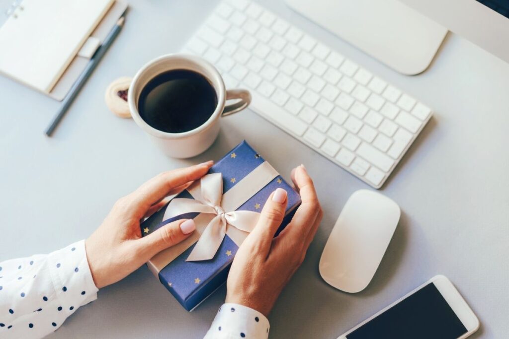 woman's hands hold a wrapped gift in front of the computer on her desk. 