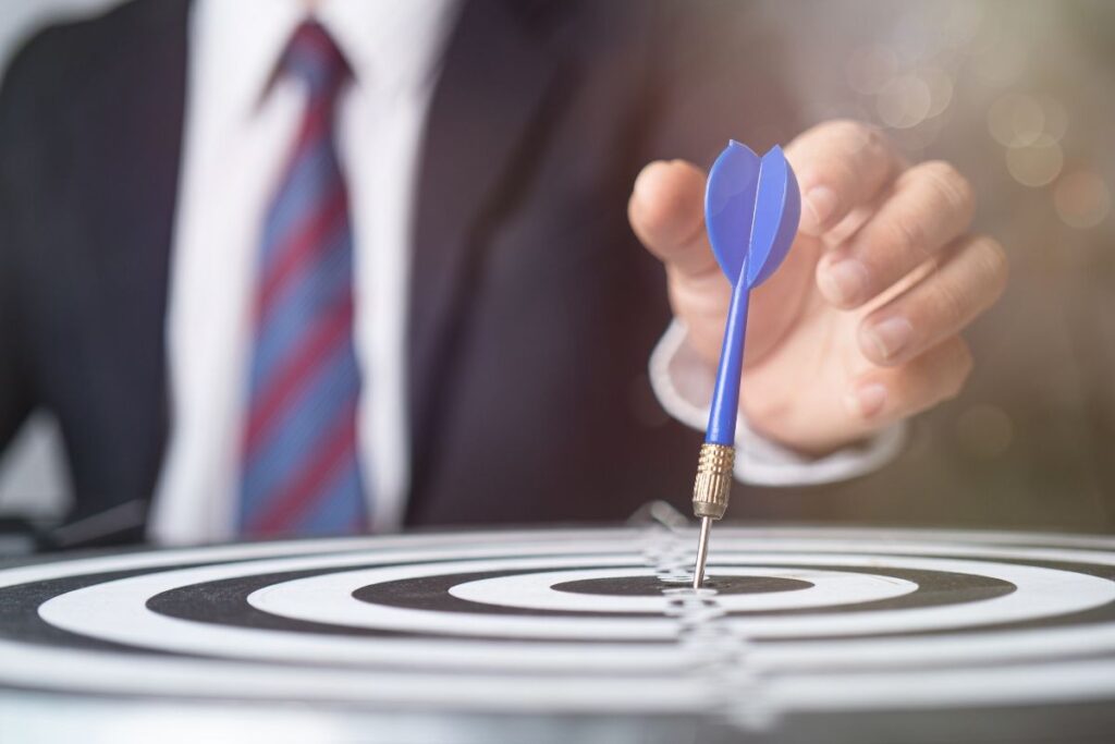 A man in a suit reaches to a dart in the centre of a dart board. 