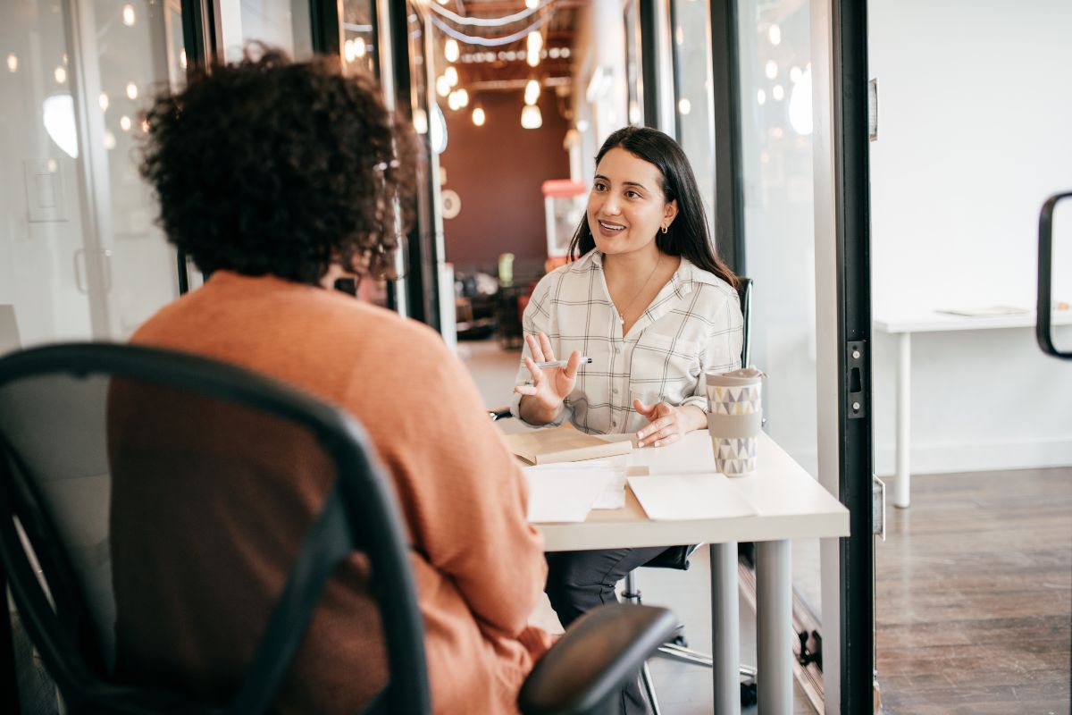 Two women sit across from each other. One interviews the other. They are in a professional office space and wear office workwear.