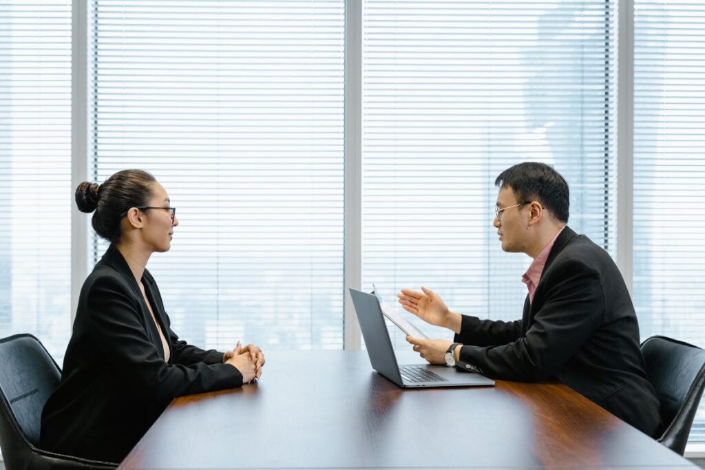 a woman and a man sit opposite each other in a professional interview. They are both wearing suits.