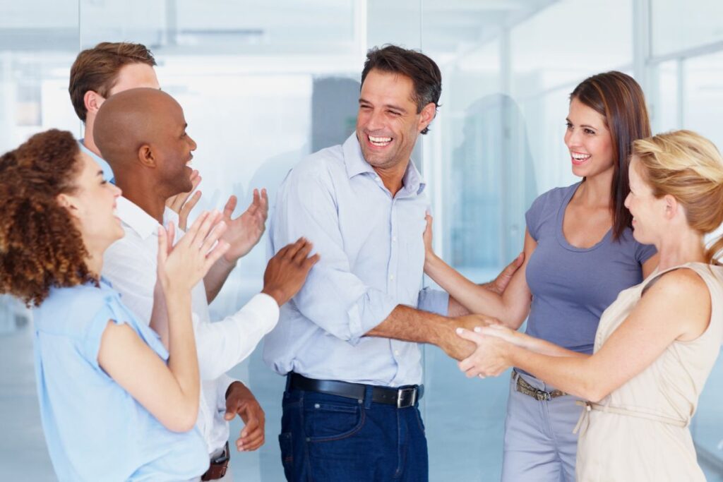 a group of colleagues stand in a circle and shake hands with a man in the centre. They are all smiling and laughing, and some are clapping in celebration.