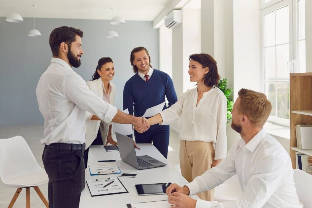 a group of colleagues gather around a table. One woman shakes hands with a man in the middle. The others smile. There are laptops and paperwork on the table in front of them. 