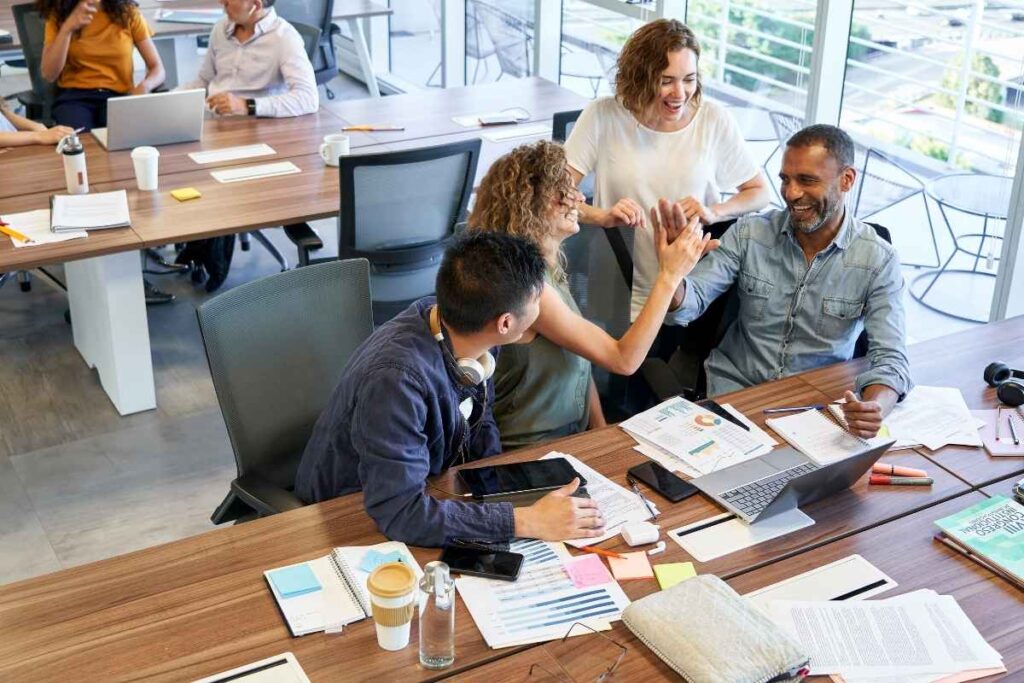 a group of 4 colleagues gather together in the office. A man and woman in the centre high five. They are all smiling. There is a laptop on the table alongside lots of paperwork. 