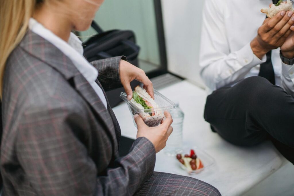 A make and female colleague sit and eat sandwiches together on their lunch break. They are both wearing suits. Their faces are not on the photo.