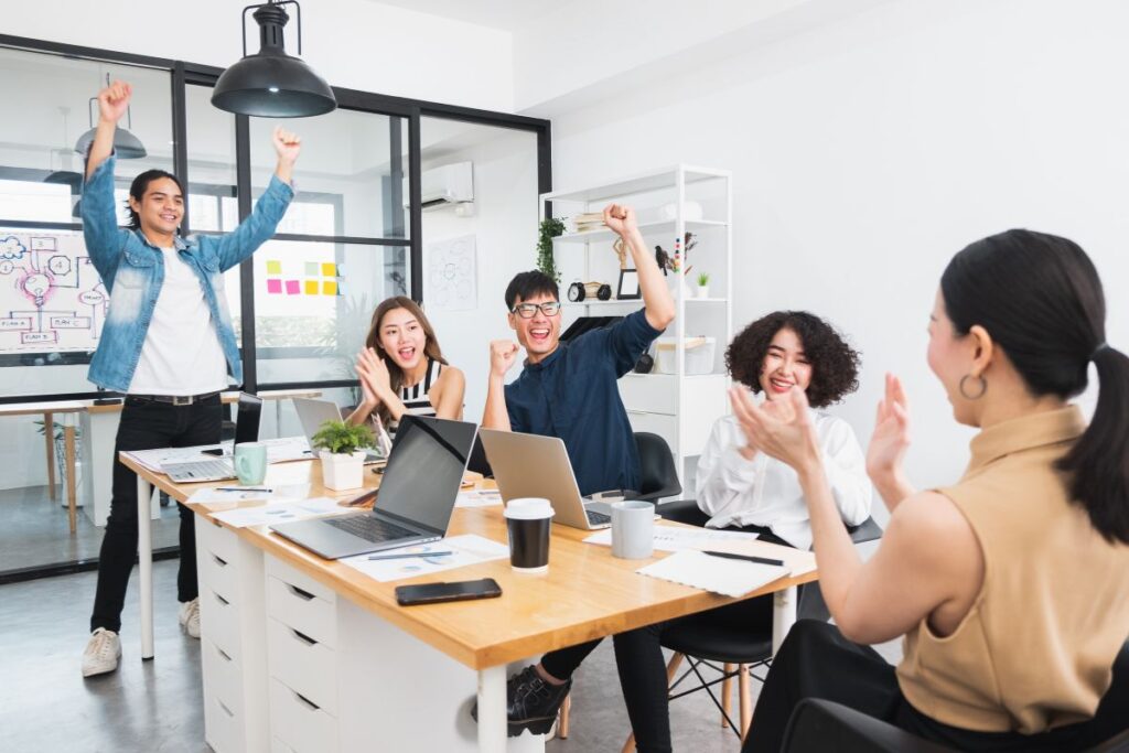 A group of colleagues celebrate together around a table. Two men hold their hands in the air whilst three women clap. There are laptops in front of them.