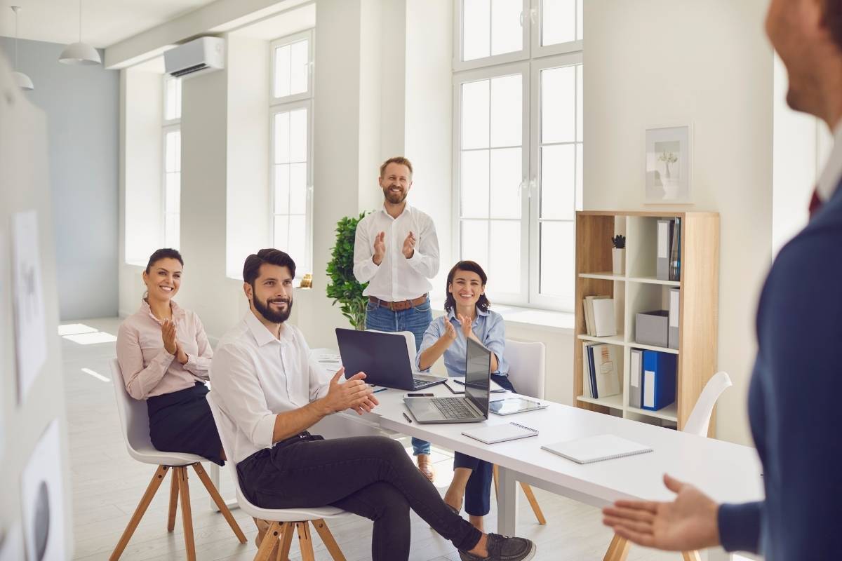 A group of colleagues sit in a group and clap and celebrate a man stood up in front of the group