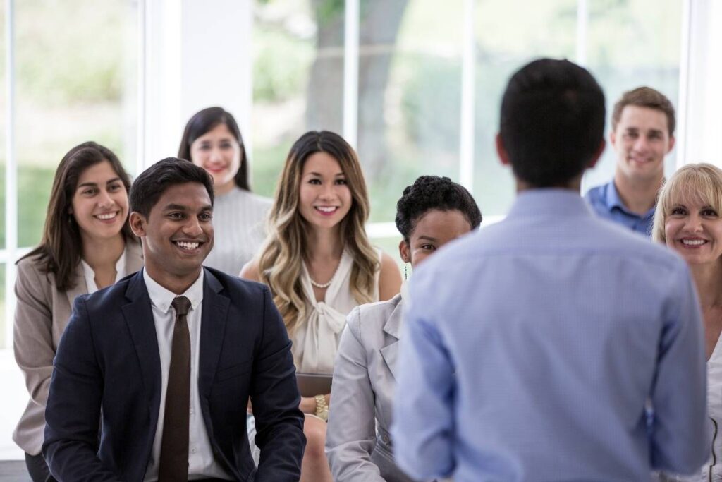 a man stands in front of his co-workers and does a presentation to them. They are all smiling.  