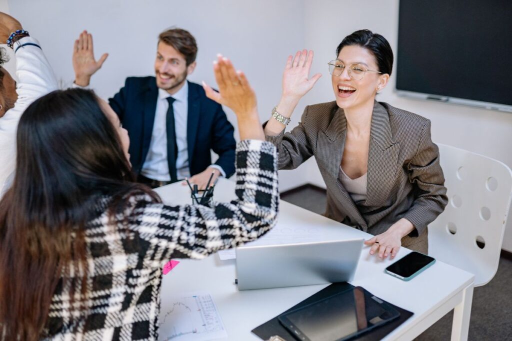 two female colleagues high five each other over a desk. In the background, two male colleagues also high five. They are all smiling.