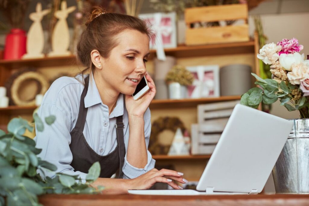 A woman sits at her work laptop whilst taking a phone call. She has flowers on her desk.