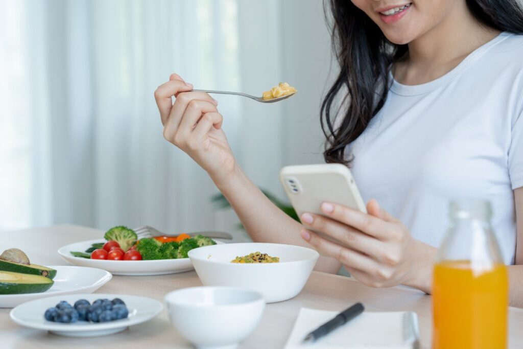 a woman eats a selection of healthy food whilst scrolling on her phone