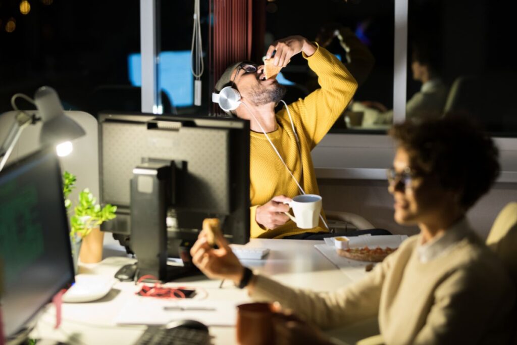 two co-workers sit at their desk at night and snack on pizzas and drink hot drinks.