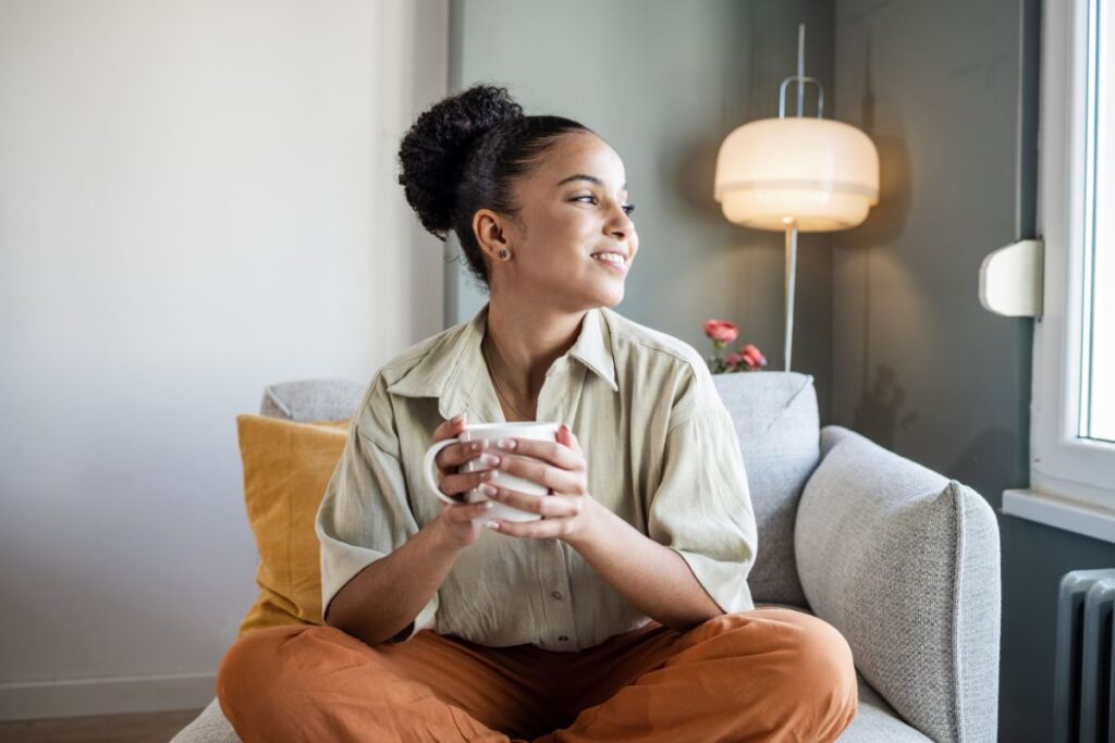 A woman sits cross legged and smiling as she stares out of the window. She is holding a mug. 