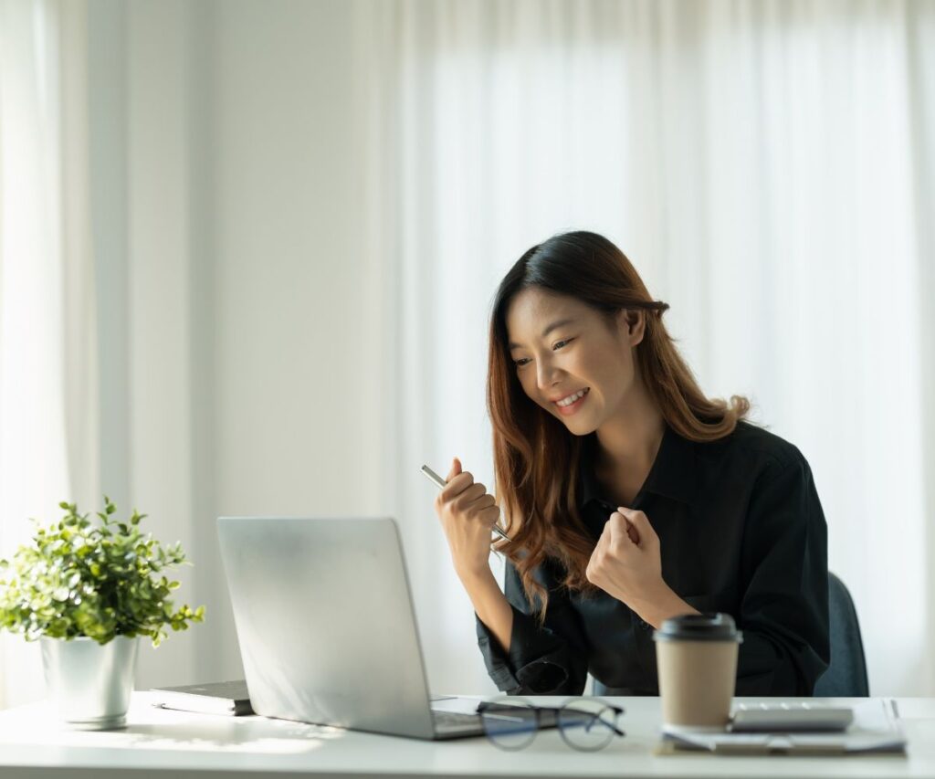 a smiling woman works at her desk. There is a laptop and a cup of coffee in front of her.