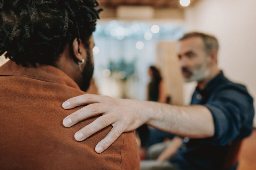 One man places his hand on another man's shoulder. The man is blurred in the background. but his hand is in focus.