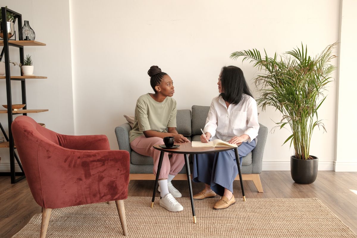 Two women sit together and offer support to each other over a coffee. One of the women is writing down the conversation.