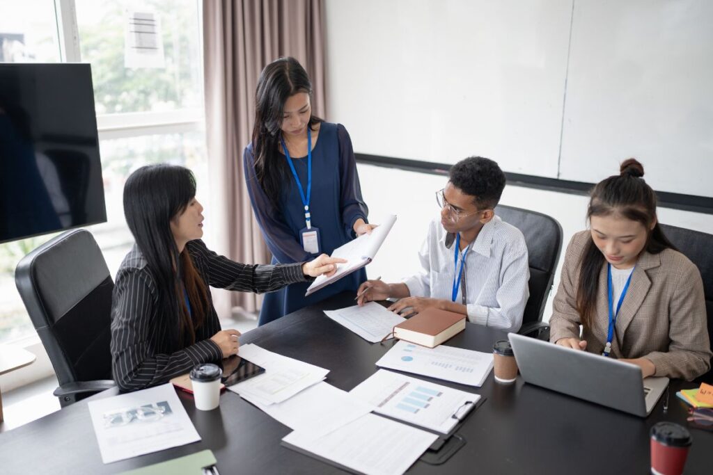 A woman stands and talks through the notes on her sheet. Three of her colleagues sit around the table and work on their laptops or make notes.