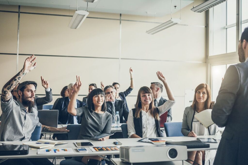 A group of employees sit together and listen to a speaker. They all have their hands raised into the air as though answering a question.