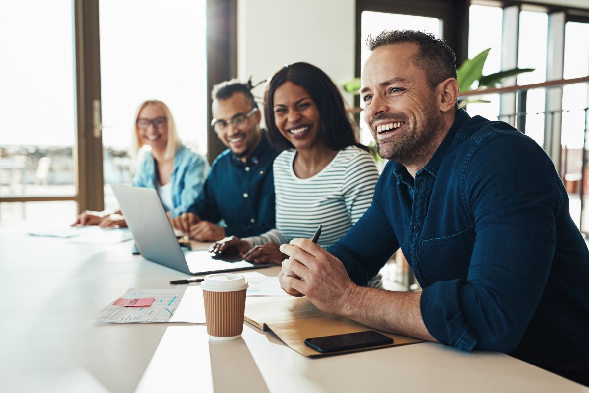 A group of co-workers sit together at work. There is a laptop and a coffee on the table
