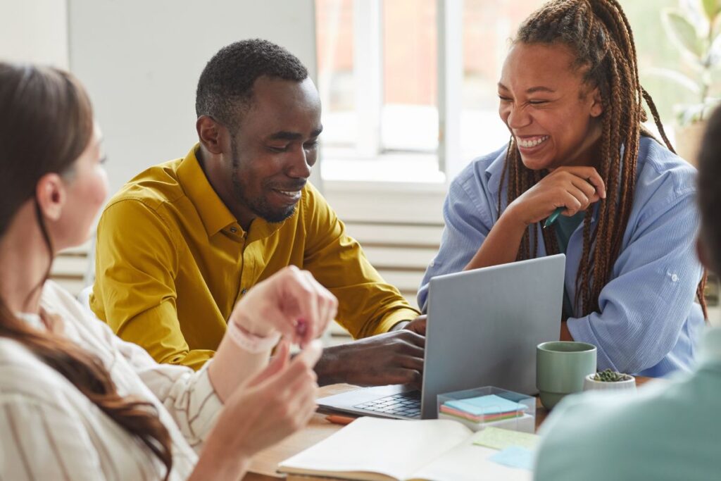 A group of co-workers sit together at a table. One woman laughs while the man next to her smiles and works on his laptop.