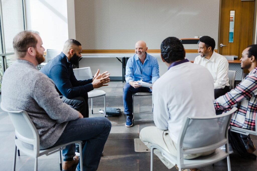A group of male colleagues sit in a support circle and smile and laugh together.
