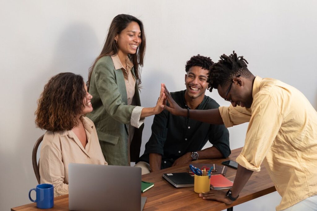 A group of four employees sit together and laugh. There is a laptop on the table and the two people in the middle high five each other over the table.