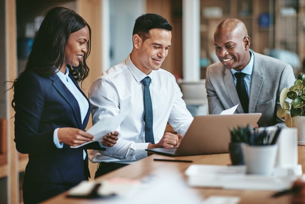 A group of three colleagues in formal workwear look at a laptop together and smile.