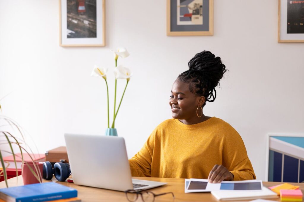 a woman sits at her desk and smiles as she works from home. There is a laptop in front of her.