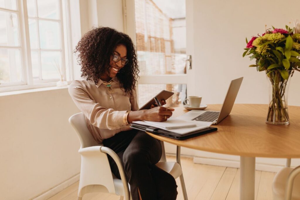 A woman sits at her desk and smiles. She writes notes and holds a calculator.