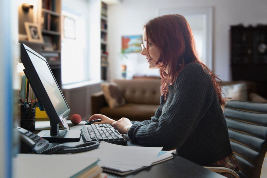 A woman sits at her desk and works on her computer. There is a sofa and a bookshelf in the background.