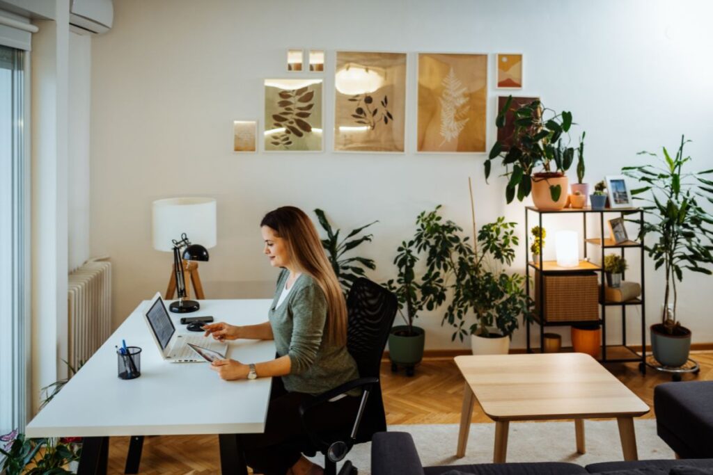 A woman sits at her desk and works at her laptop. She is holding a tablet in one hand.