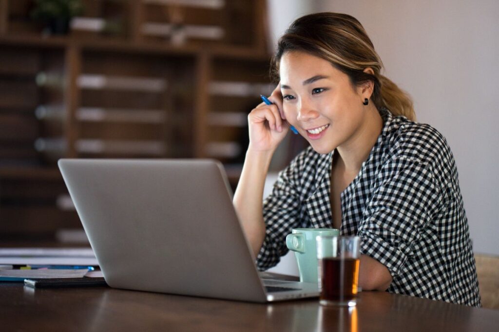 A woman sits at her desk and smiles. She is looking at her laptop and there is a cup of coffee in front of her.