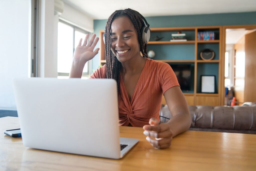 A woman sits on a remote meeting at home. She smiles and waves at the camera. She is wearing headphones.