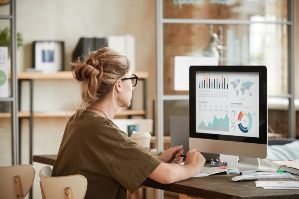 A woman sits on her computer and reads data. She has her hair in a bun and is wearing glasses.