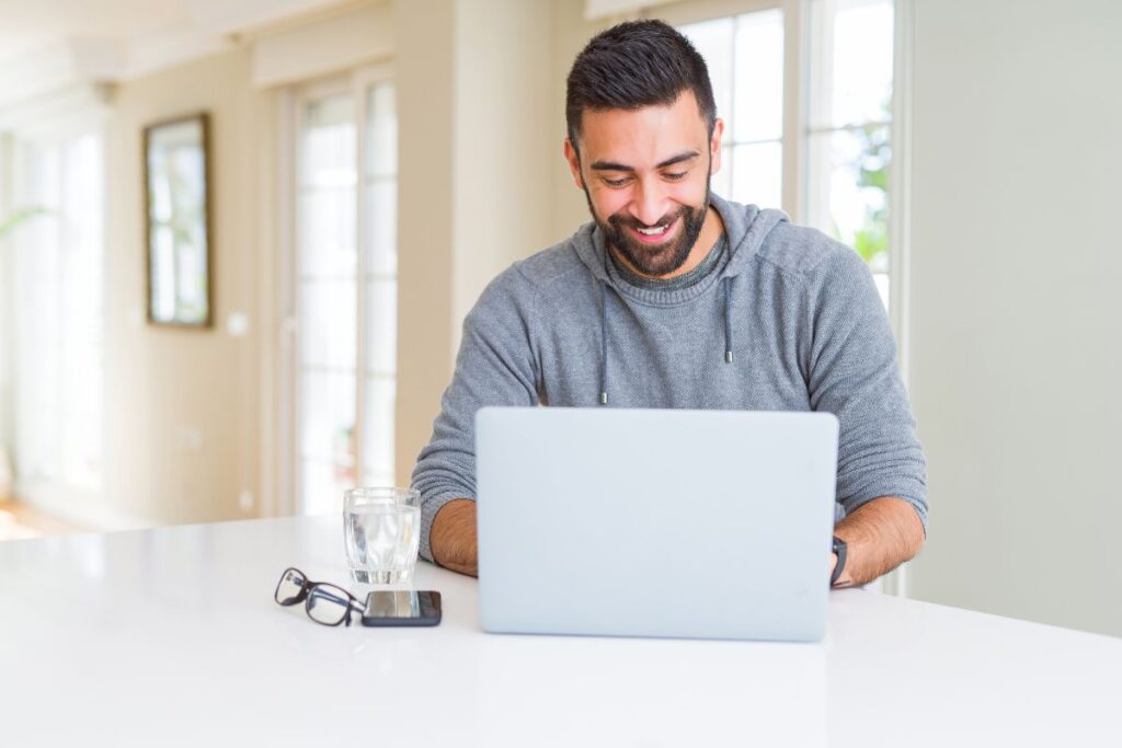 A man sits on his laptop and smiles. There is a glass of water on the table, alongside his glasses and phone.