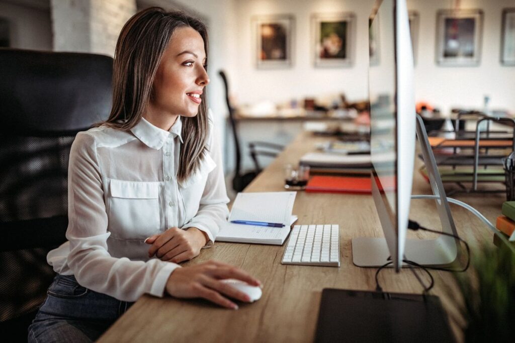 A woman sits on her computer and scrolls. She is wearing a white shirt and smiling.