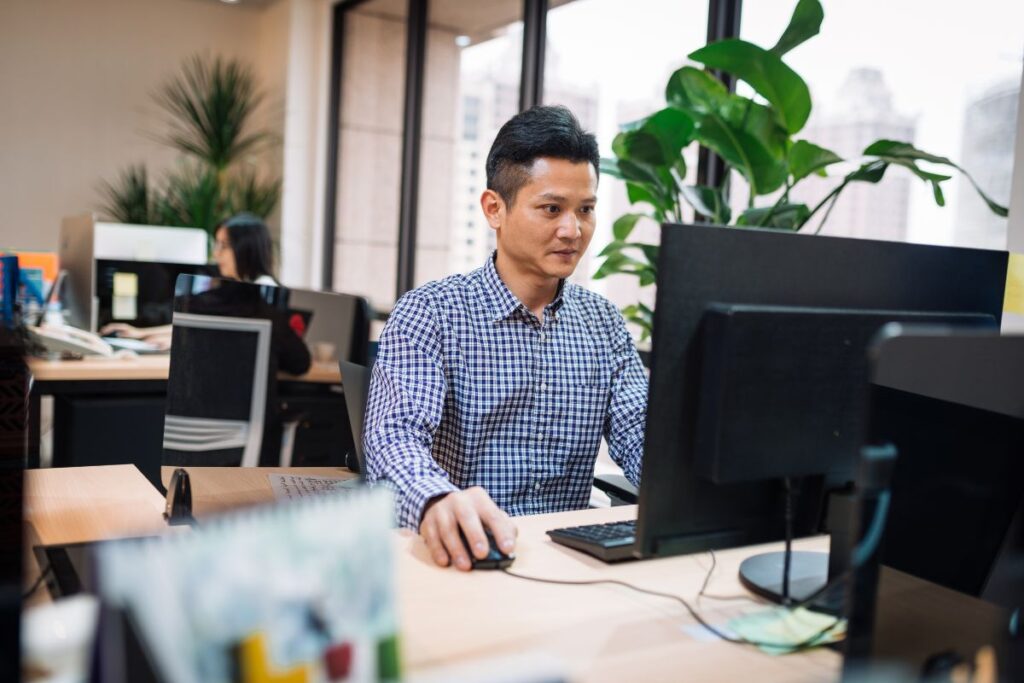 A man sits on his computer at work. In the background of the photo, you can see the rest of the office.